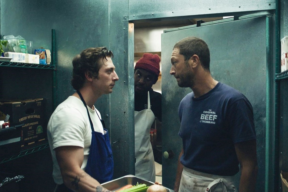 The image captures a tense moment from the TV show "The Bear," set in the high-pressure environment of a restaurant kitchen. In a cramped space, three characters are engaged in a serious discussion. One, wearing a white shirt and blue apron, looks intensely focused, suggesting a mix of determination and frustration. The second character, peeking from behind, adds an element of curiosity or mischief, while the third, dressed in a dark shirt, appears contemplative, possibly weighing the gravity of the conversation.
The metallic backdrop and clutter of kitchen supplies accentuate the chaotic atmosphere typical of restaurant life. The close quarters symbolize the pressure of culinary art, where every second matters. This scene humorously highlights the absurdity of intense kitchen dynamics, as the characters navigate their roles amidst the chaos, making it relatable to anyone who's ever felt overwhelmed in a high-stakes situation.