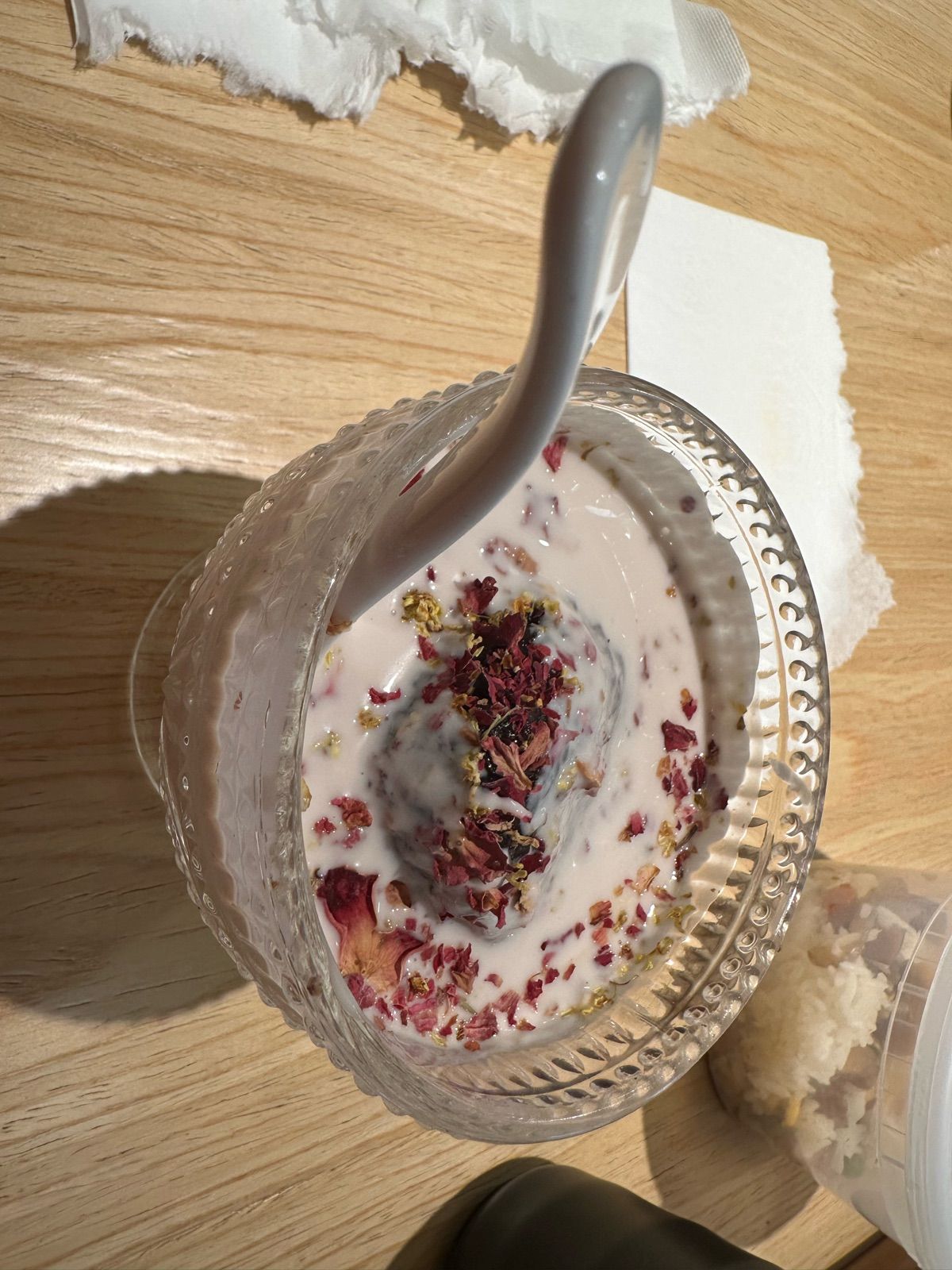 This overhead shot captures a decadent dessert, a dark, mysterious scoop nestled in a creamy, pale liquid within a textured glass bowl. The dessert, seemingly a shy yet flamboyant character, is adorned with a lavish 'outfit' of dried red rose petals and golden sprinkles, giving it a regal, slightly dramatic demeanor. A sleek white spoon, poised for a delightful plunge, hints at the imminent indulgence. The wooden table background provides a warm, inviting stage, while a crumpled napkin and a container of extra toppings suggest a casual, yet elegant, culinary affair. The dessert seems to be saying, 'Look at me, I'm fancy, but don't be afraid to dig in!' It's a delightful, floral-infused treat, ready for its close-up and a good laugh.