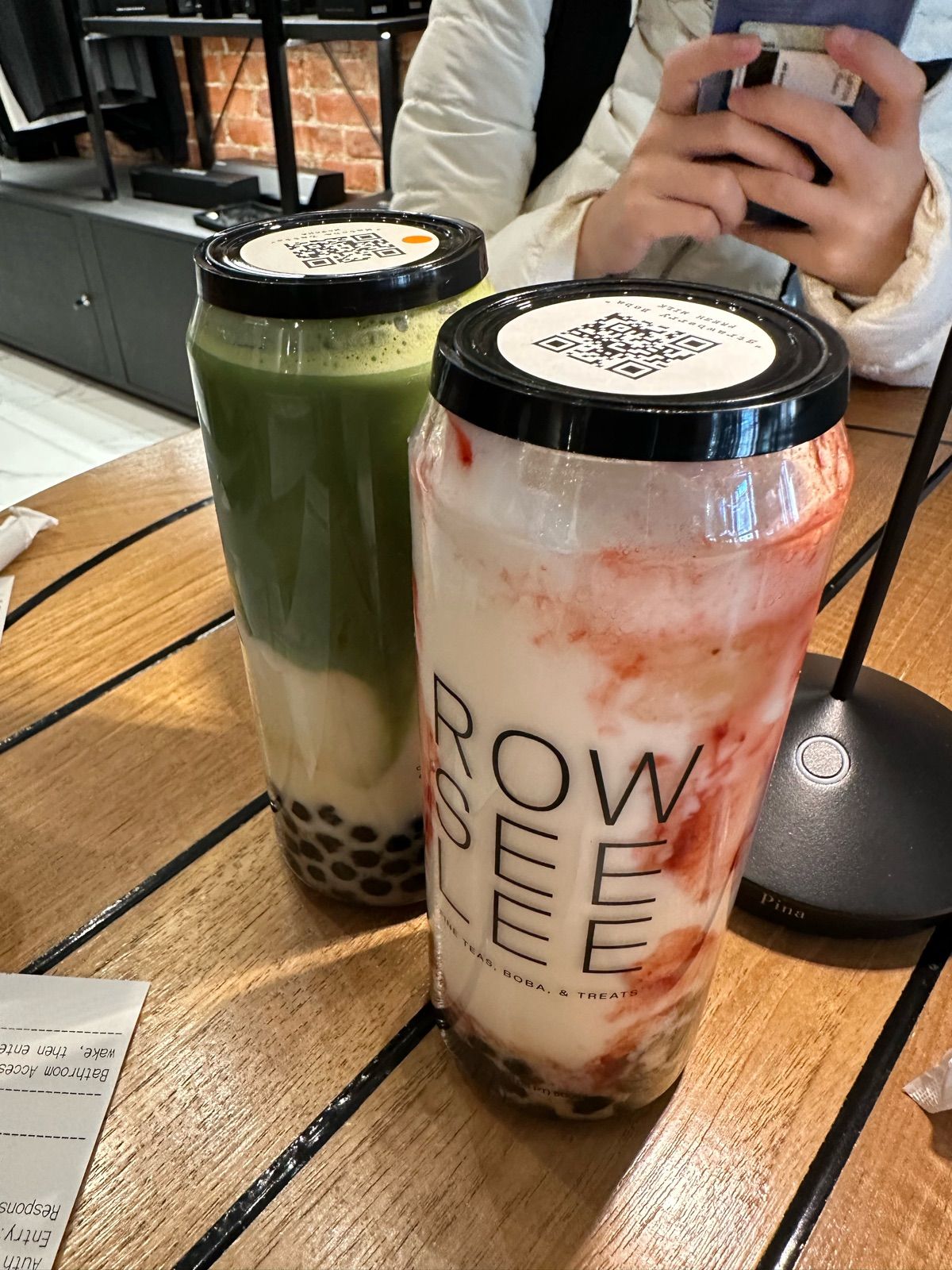 Two vibrant boba drinks, a stoic green matcha and a flamboyant strawberry swirl, stand proudly on a wooden table. The green one, layered with creamy white and dark boba pearls, seems to be the wise elder, while its red-streaked companion, also with boba, exudes playful energy. Both wear QR code 'hats,' perhaps ready to be scanned into a digital boba hall of fame. In the background, a person in a white jacket is engrossed in their phone, oblivious to the drinks' silent contemplation in this cozy cafe setting. The brick wall and dark shelving suggest a modern, relaxed atmosphere, perfectly complementing these liquid personalities awaiting their delicious destiny. I did not find any famous or recognizable content in this image.