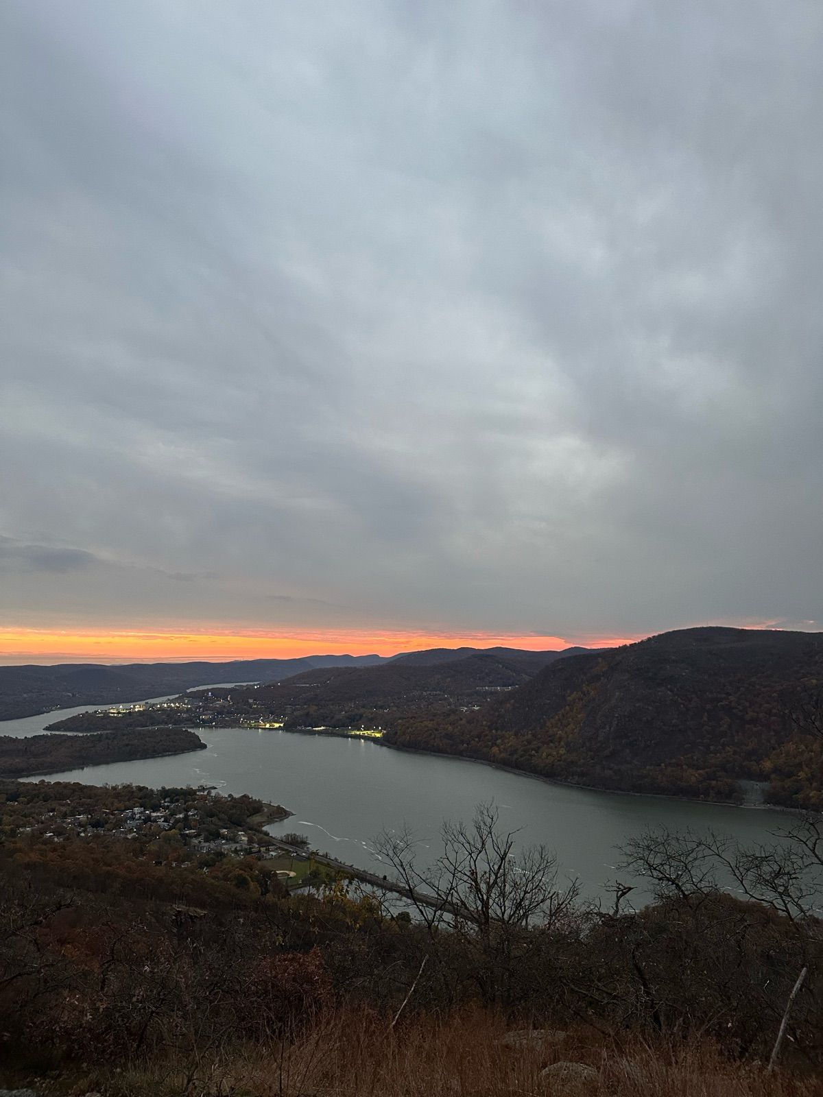 This breathtaking panoramic view captures a serene river winding through rolling hills at dusk. The sky, predominantly overcast with soft grey clouds, dramatically parts at the horizon to reveal a vibrant band of orange and pink, signaling the day's gentle farewell. Along the riverbanks, a town twinkles with numerous lights, a testament to human presence nestled within the vast natural landscape. The foreground, featuring sparse, bare branches and dry grasses, suggests a high vantage point, perhaps a quiet overlook. The landscape itself seems to be in a contemplative mood, observing the transition from day to night. It's as if the river is gracefully showing off its path, while the moody clouds reluctantly share a glimpse of the sunset's fiery personality. No famous content is recognized, just nature's quiet grandeur.