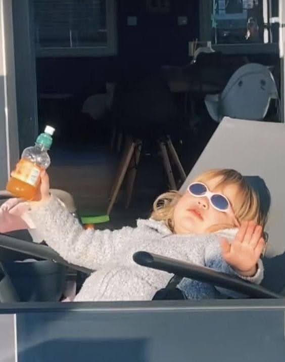A young child, likely a toddler with blonde hair, exudes an air of effortless cool, lounging in what appears to be a stroller or high chair. Donning stylish white oval sunglasses, they hold a small bottle of orange juice in one hand while raising the other in a playful 'talk to the hand' gesture. Dressed in a cozy, light-colored sweater, the child's demeanor is one of utter relaxation and sass, as if enjoying a luxurious sun-drenched holiday. The brightly lit foreground contrasts sharply with the dimly lit indoor background, visible through a doorway, featuring dark furniture and a white decorative object. This juxtaposition humorously emphasizes the child's 'vacation mode' attitude, seemingly oblivious to the mundane world behind them, embodying a tiny, sun-worshipping boss.