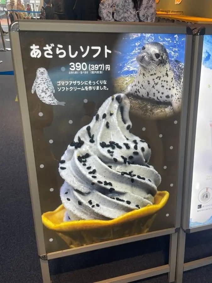This image captures an advertisement for 'Seal Soft Serve,' a delightful themed ice cream product, likely found at a Japanese aquarium or zoo. The main subject is a large, vibrant image of a spotted seal, looking directly at the viewer with a charming, almost mischievous demeanor, as if proudly showcasing its edible twin. Below it, the soft serve itself is a swirl of greyish-white ice cream, generously speckled with black sesame seeds to perfectly mimic the seal's distinctive spots, served in a bright yellow waffle cone. A smaller, cartoon-like seal also graces the ad. The blurred background of people suggests a bustling public environment, reinforcing the context of a popular attraction. The humor lies in the adorable seal seemingly endorsing its own delicious, spot-on dessert doppelgänger, a playful nod to Japan's love for cute, themed foods.