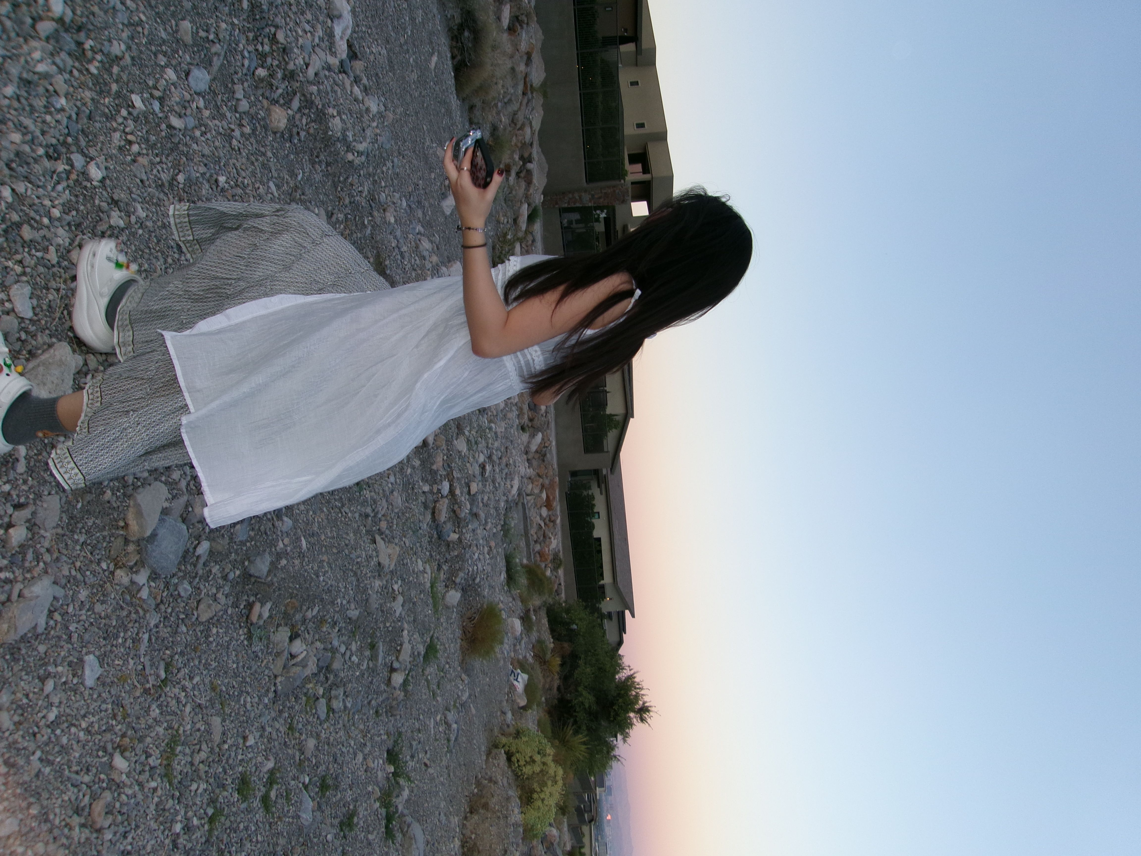 This image captures a person from behind, their long dark hair flowing, as they seemingly attempt to photograph a scenic view. Dressed in a flowing white tunic over patterned pants and white Crocs adorned with colorful charms, they hold both a silver digital camera and a smartphone, perhaps debating which device will best immortalize the moment. The subject's casual attire suggests a relaxed, unpretentious demeanor, focused on capturing the beauty before them. The background reveals a rugged, gravelly terrain leading up to modern, multi-story buildings with dark windows, possibly a resort or residential complex, set against a vast sky transitioning from soft blue to a warm pinkish-orange, indicative of dusk or dawn. The juxtaposition of the natural, rocky landscape with contemporary architecture creates an interesting visual dynamic. The entire scene is humorously tilted, making it appear as if the world itself has decided to lean into the shot, or the photographer is simply too cool for conventional angles.