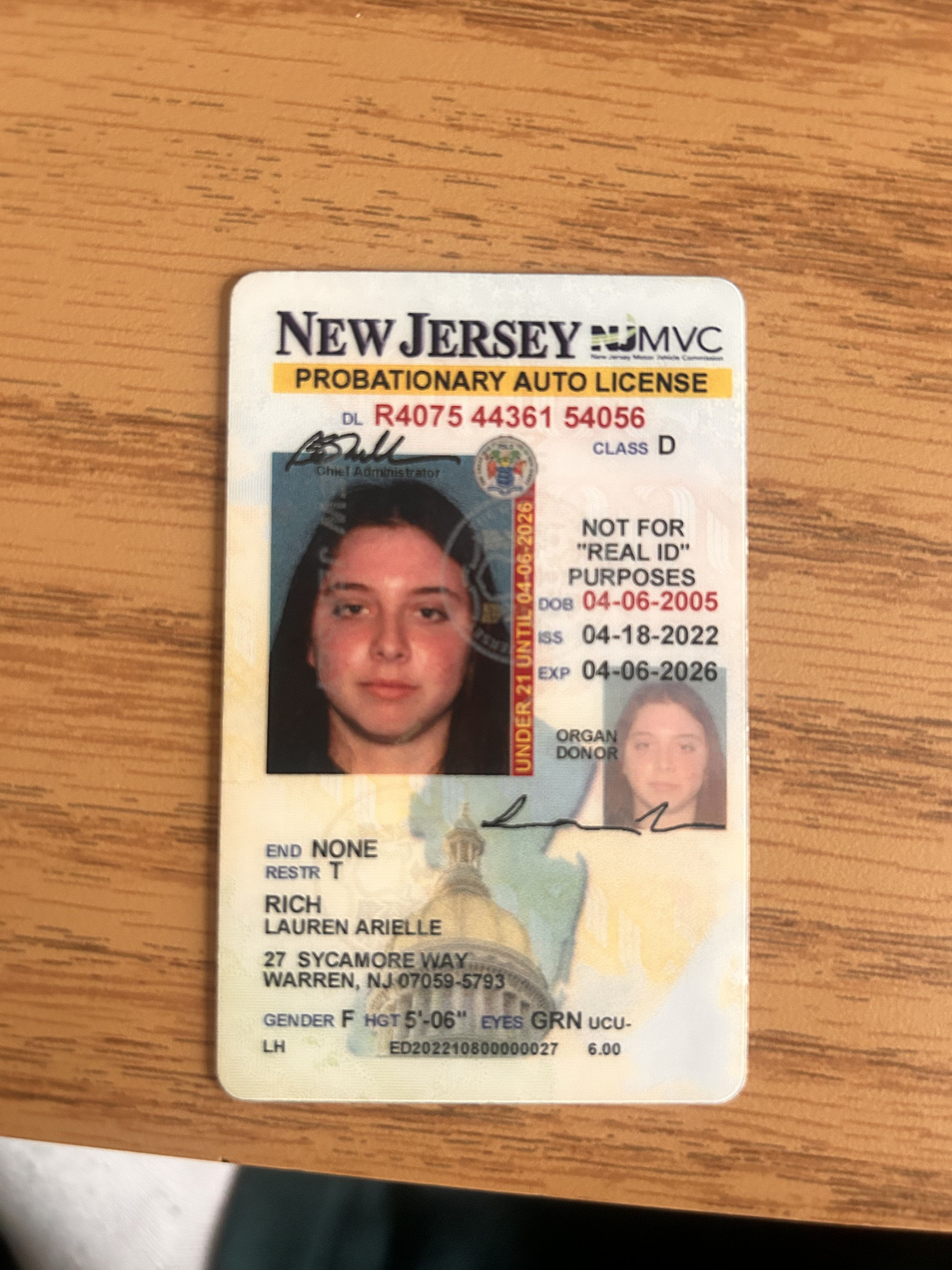 This image prominently features a New Jersey Probationary Auto License for Lauren Arielle Rich, resting on a light brown wooden surface. Lauren's main photo depicts a young woman with long dark hair and a neutral, calm expression, typical for an official document, conveying a serious demeanor. The license's background subtly incorporates the New Jersey State House dome, linking the document to the state's identity. Humorous elements include the bold red "UNDER 21 UNTIL 04-06-2026" bar, playfully emphasizing her youth and the restrictions of a probationary license. The "NOT FOR 'REAL ID' PURPOSES" text also adds a touch of bureaucratic irony, given it's a real identification document.