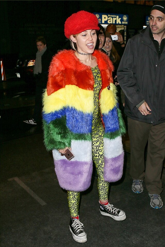 Miley Cyrus makes a vibrant public appearance, exuding energetic confidence with an open-mouthed expression, possibly singing or speaking to onlookers. She's dressed in an eye-catching, multi-colored rainbow faux fur coat, paired with bright yellow leggings adorned with a distinctive black smiley face motif, a symbol often associated with her "Happy Hippie Foundation." A fluffy red beret and classic black Converse high-top sneakers complete her eccentric ensemble. She holds an iPhone in her left hand. The background, featuring a "Park" sign and several people, including one holding a phone to photograph her, clearly indicates a public event where she is the center of attention. The humorous clash of textures and patterns highlights her unique fashion sense.