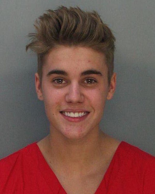 This image is a widely recognized mugshot of pop superstar Justin Bieber, taken during his 2014 arrest in Miami Beach. He is seen smiling broadly, almost gleefully, directly at the camera, a demeanor strikingly unusual for a booking photo. His light brown hair is styled in a tousled, spiky fashion, and he wears a bright red garment, likely standard jail attire. The background is a plain, light grey wall, typical of a police station booking area, which starkly highlights his cheerful expression. The humor in the image stems from the stark contrast between the serious context of an arrest and Bieber's seemingly unbothered, almost triumphant smile, making it an iconic meme.