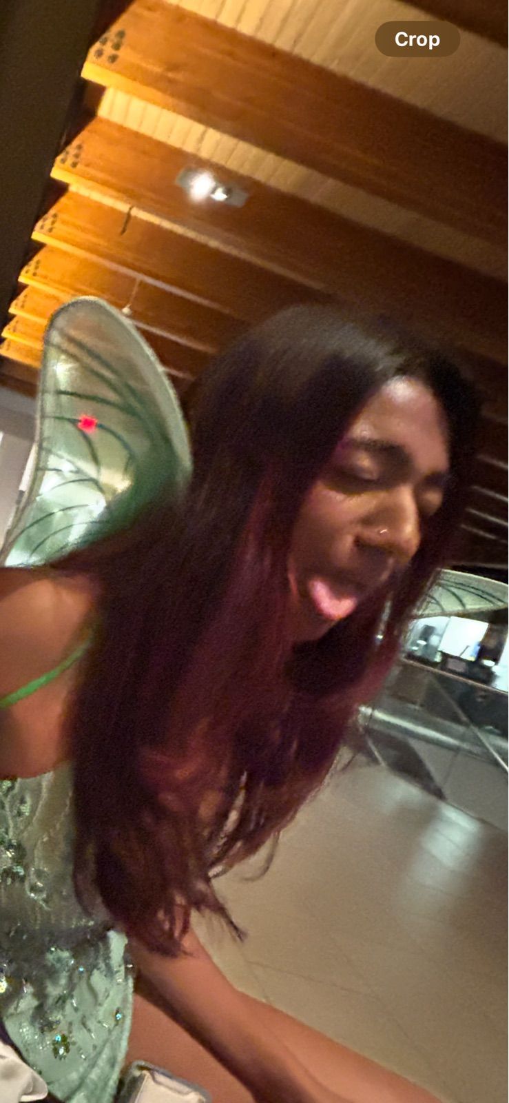 A person with long, dark hair, possibly with reddish undertones, is captured mid-pose, playfully sticking out their tongue with eyes partially closed. They are wearing a light green, possibly sequined, top with a thin strap and prominent, translucent green fairy wings, suggesting a costume or themed event. A small nose piercing is visible. The subject's demeanor is clearly lighthearted and mischievous. The background features warm wooden ceiling beams, indicating an indoor setting, possibly a cozy restaurant or event space, which complements the festive attire. The overall image conveys a fun, candid moment, with the cheeky expression adding a humorous touch to the fairy ensemble.