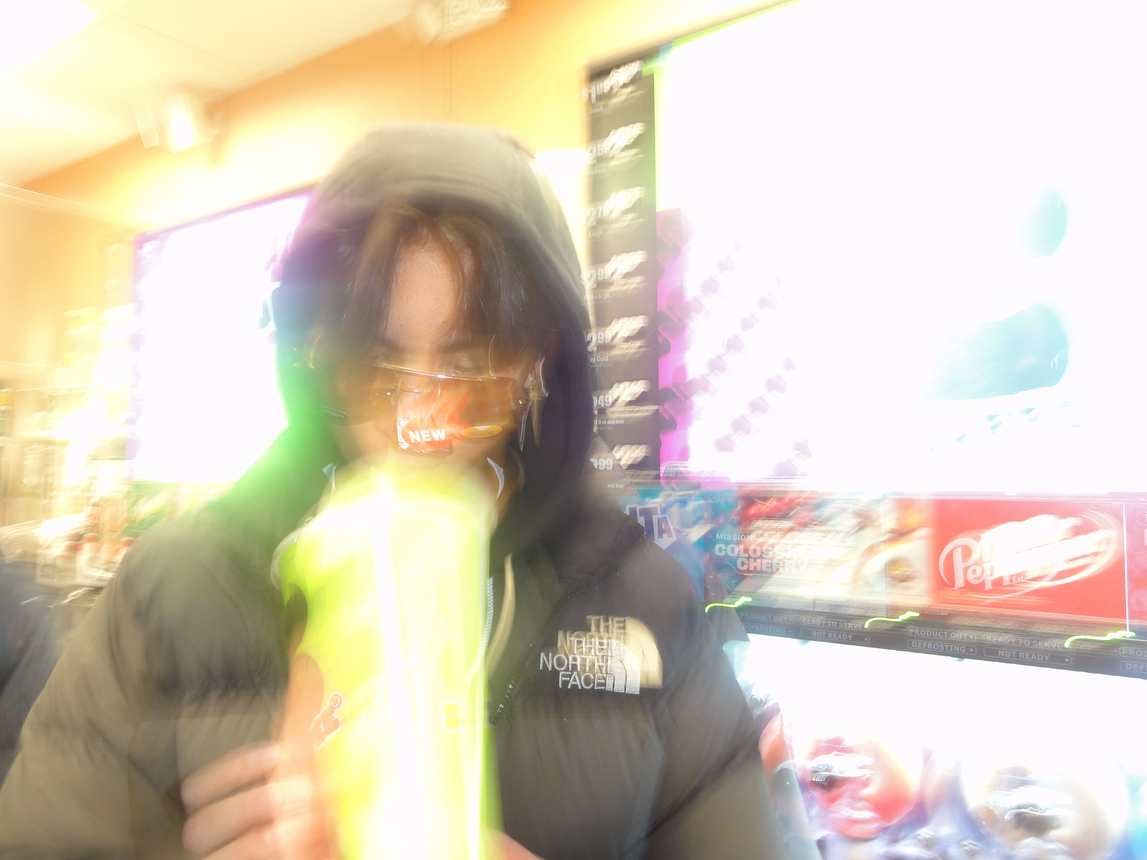 The image features a person dressed in a puffy black jacket and a hood, holding a large, brightly colored beverage. Their demeanor suggests a casual, perhaps playful attitude, as they seem to be mid-action, possibly taking a sip or preparing to do so. The blurred effect around them gives a sense of motion, enhancing the animated atmosphere of the scene.  

The background reveals a convenience store setting with vibrant advertisements and product displays, hinting at a bustling environment. The bright colors and illuminated signs create a lively backdrop, suggesting a late-night snack run or a fun outing with friends. The overall composition evokes a sense of spontaneity and youthfulness, reminiscent of casual hangouts or relatable moments captured in popular culture.