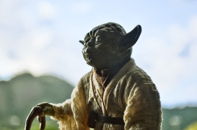 This close-up image features a highly detailed action figure of Yoda from Star Wars, captured in a contemplative pose. His iconic green skin, large pointed ears, and wise, slightly puckered expression are prominent. He wears his traditional tan and brown Jedi robes, appearing weathered, and his gnarled cane is visible in his left hand, suggesting his advanced age and reliance on it. The background is a softly blurred outdoor scene, with distant green hills or mountains under a bright, slightly overcast sky. This natural setting evokes the serene, yet wild, environments often associated with Yoda, such as Dagobah, where he trained Luke Skywalker. The image humorously portrays the venerable Jedi Master seemingly enjoying a peaceful moment, perhaps pondering the Force or simply appreciating a nice day, far from galactic conflicts.