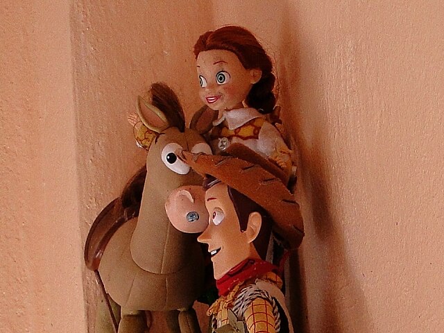 This image presents a charming and humorous display of beloved characters from the Toy Story franchise: Jessie, Bullseye, and Woody, depicted as detailed figurines. Jessie, with her wide-eyed enthusiasm and beaming smile, is playfully perched atop Bullseye, who looks equally eager with perked ears and a joyful expression. Below them, Woody, sporting his iconic cowboy hat and a classic friendly grin, forms the sturdy base for this whimsical pile-up. The characters exude a cheerful and adventurous demeanor, as if caught mid-action in one of their escapades. The plain, light brown wall in the background serves as a simple, unobtrusive backdrop, allowing the vibrant personalities of these iconic toys to take center stage. The humorous stacking suggests a moment of playful camaraderie, perfectly encapsulating the spirit of friendship from the films.