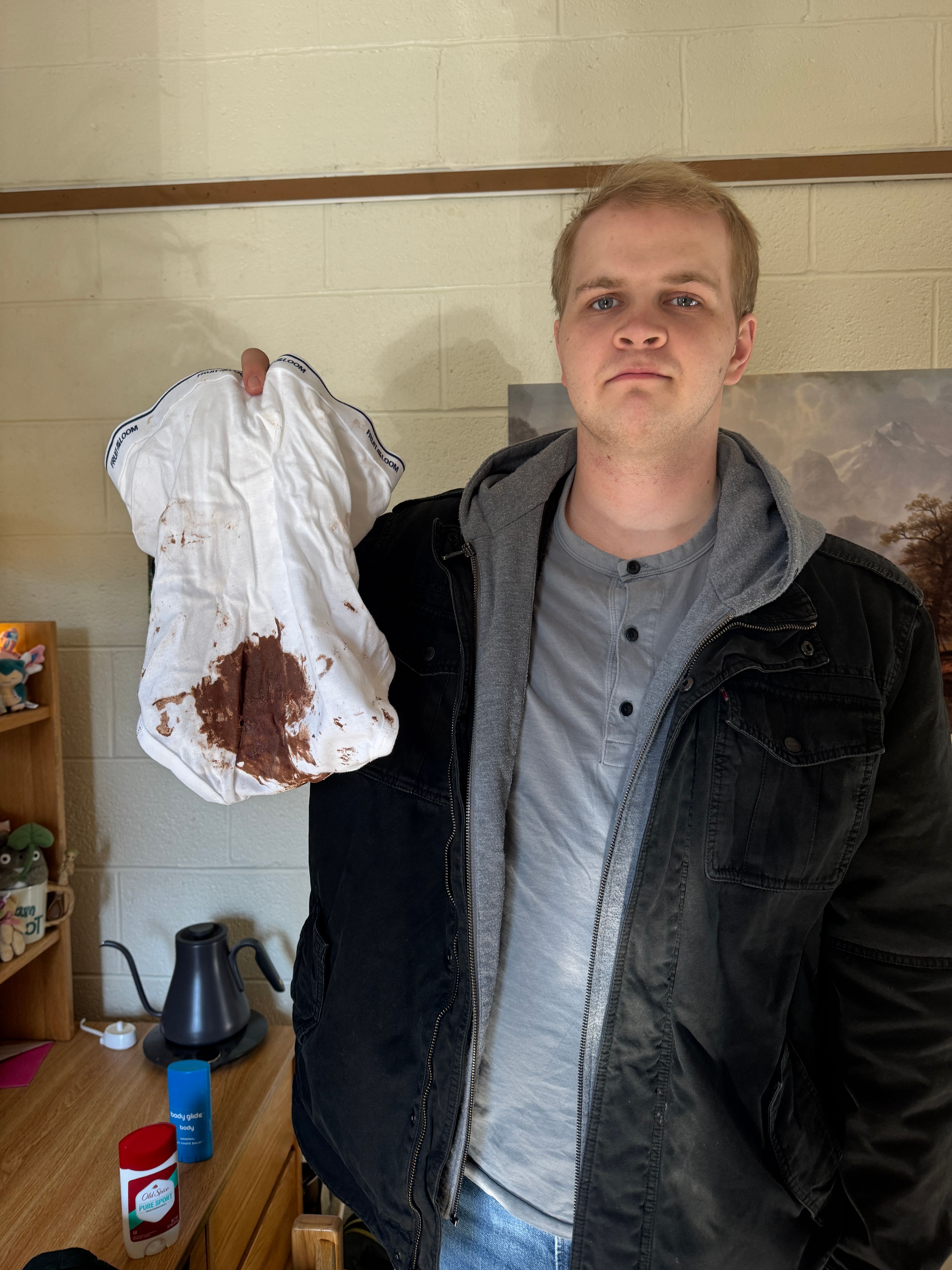 In the image, a young man stands with a serious expression, holding up a pair of white shorts stained with what appears to be brown substance, possibly chocolate or mud. His demeanor suggests a mix of annoyance and bemusement, as if he’s just discovered an unfortunate mishap. He is dressed in a black jacket over a gray shirt, contrasting with the casual nature of the shorts he holds.
The background features a simple room, with a wooden table displaying various items including a bottle and a container, giving it a cozy yet cluttered feel. The light is bright, suggesting daytime, and the overall atmosphere feels humorous, as if he’s caught in a comedic moment of domestic chaos. The image evokes a relatable scenario where one might accidentally ruin clothing, highlighting the humorous side of everyday life.