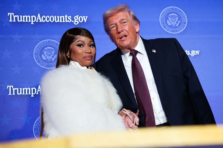 This image features rapper Nicki Minaj and former President Donald Trump in a public appearance. Minaj, in a lavish white fur coat, gazes directly at the camera with a somewhat serious, slightly pursed expression. Trump, in a dark suit, appears mid-sentence with an animated, open-mouthed look. Their hands are clasped, creating an unusual formality. The blue background, emblazoned with "TrumpAccounts.gov" and the Presidential Seal, firmly places this event within a political context related to Trump's administration, likely a White House visit. The juxtaposition of the glamorous artist and the former president in this official setting creates a humorous, almost surreal visual, highlighting the unexpected nature of their interaction.
