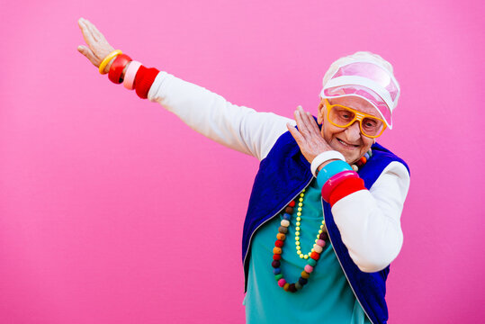 The image features an elderly woman striking a playful pose, confidently executing the 'dab' dance move, which adds a humorous touch to the scene. Her vibrant outfit, adorned with colorful bangles and a bright hat, suggests a fun-loving personality, radiating joy and lightheartedness. The backdrop is a bold pink, amplifying the cheerful vibe and enhancing the playful context of the image.
This scene embodies a current trend in social media culture, where age is no barrier to having fun and embracing youthful expressions. The woman’s demeanor is lively and spirited, showcasing a delightful contrast to conventional expectations of aging. Overall, the image captures a moment of joy, reminding viewers that laughter and playfulness can thrive at any age.