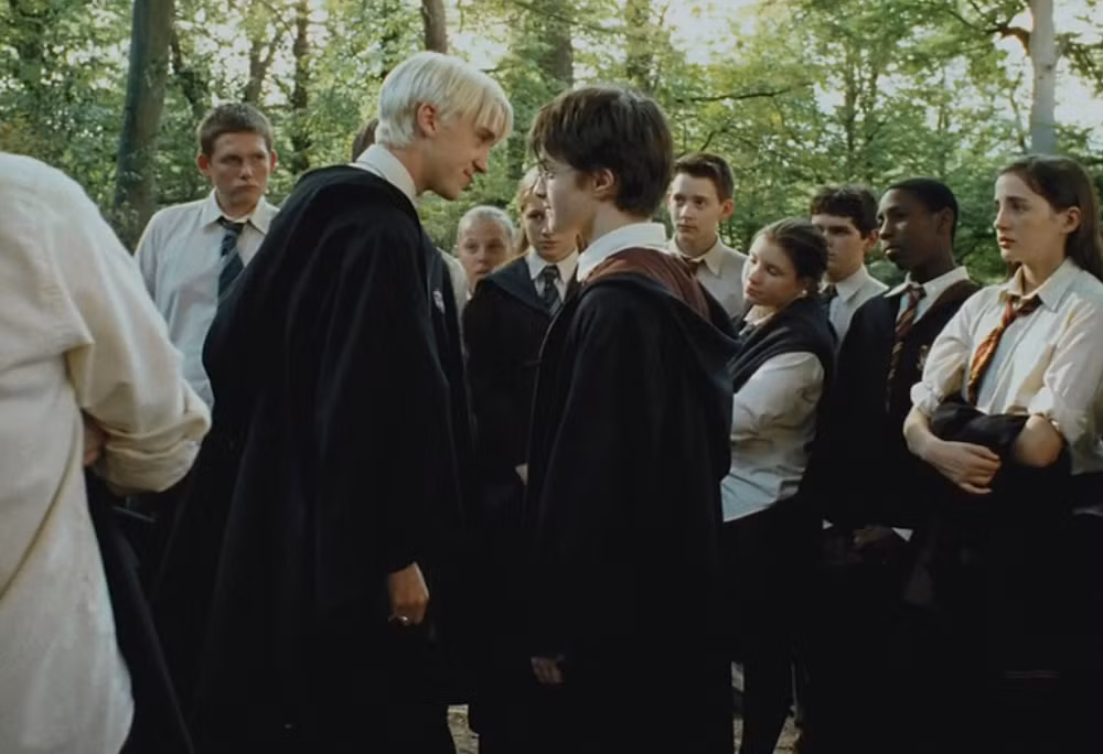 This image captures a tense moment from the Harry Potter film series, specifically featuring Daniel Radcliffe as Harry Potter and Tom Felton as Draco Malfoy in a confrontational stare-down. Draco, with his signature platinum blonde hair, leans in with a sneering, almost amused expression, while Harry, in his round glasses, meets his gaze with a serious, unyielding demeanor. Both are clad in their Hogwarts robes, indicating they are students at the School of Witchcraft and Wizardry. Surrounding them, other students observe the unfolding drama with varied expressions of curiosity and concern, creating a palpable sense of anticipation. The background, a lush, sun-dappled wooded area, perfectly sets the scene for a typical outdoor confrontation on the Hogwarts grounds. The humor lies in the classic, almost theatrical rivalry between the two, with an entire audience of students seemingly enjoying the impromptu 'show'.