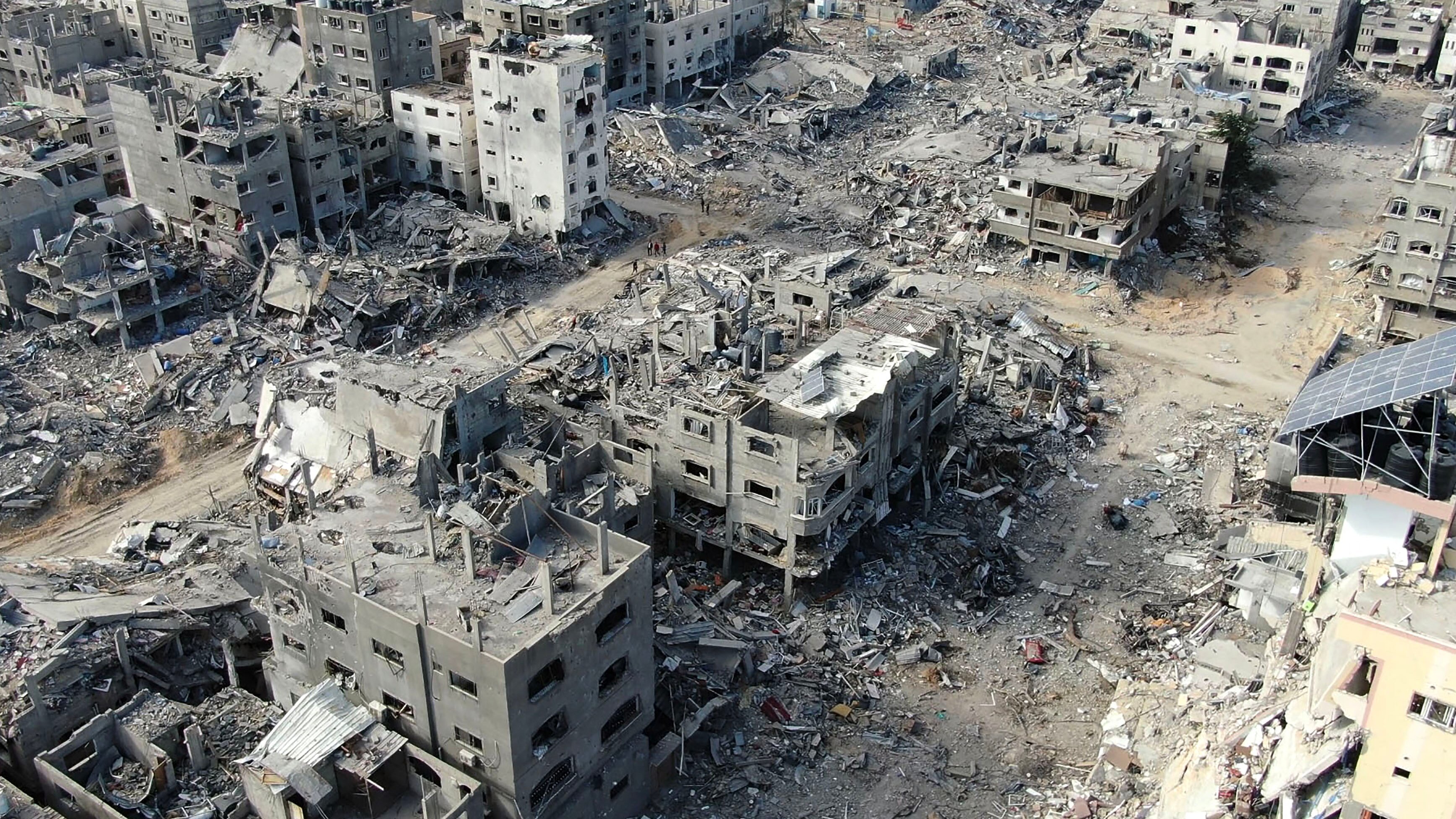 The image depicts a desolate urban landscape filled with the remnants of collapsed buildings and debris, indicative of significant destruction, likely due to conflict or disaster. The structures are mostly gray and crumbling, with exposed interiors and scattered rubble creating a stark, somber atmosphere.
In the foreground, a few people can be seen navigating through the wreckage, their small figures emphasizing the vastness of the devastation. The background features additional ruined buildings, creating a sense of disarray and hopelessness.
This scene, devoid of life and vibrancy, evokes a feeling of loss and despair. The absence of greenery or signs of recovery underscores the dire state of the area. Overall, the image powerfully conveys the consequences of human conflict, serving as a haunting reminder of the fragility of urban life.