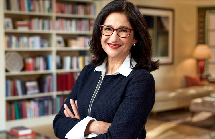 The image features a confident woman with a warm smile, dressed in a smart black outfit with a white collar. Her glasses add a touch of sophistication, and her posture—arms crossed and leaning slightly forward—exudes both approachability and professionalism.
The background reveals a cozy, well-organized room filled with bookshelves, suggesting a cultured environment, possibly a home office or a study. A hint of soft lighting from a lamp enhances the welcoming atmosphere. The presence of books indicates a love for knowledge and learning, further accentuating her intellectual demeanor.
This scene captures a moment of poised self-assurance, perhaps indicative of a successful professional or thought leader. The whole image gives off an inviting yet authoritative vibe, making one wonder what insights or stories she might share. It’s like she’s ready to enlighten you while still making you feel at home!