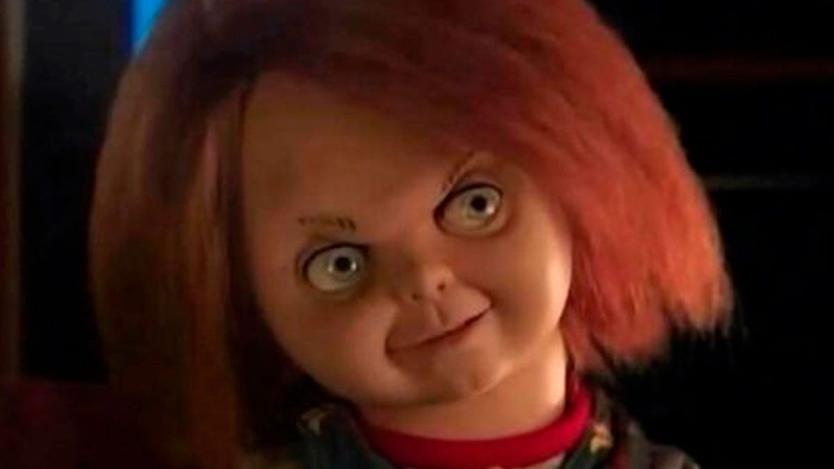 The image presents a close-up of Chucky, the iconic killer doll from the "Child's Play" horror franchise. His bright red, shaggy hair frames a pale, plastic face with wide, piercing blue eyes that stare intently. A mischievous, almost sinister grin stretches across his face, conveying a demeanor of pure, unadulterated wickedness, as if he's just conceived a truly diabolical plan. He wears a red collar, hinting at his signature overalls. The background is dark and indistinct, suggesting a dimly lit, ominous setting that perfectly aligns with his terrifying persona. The humor stems from the doll's exaggerated, almost cartoonish expression of evil, making him look like a mischievous toddler who's about to cause delightful chaos.