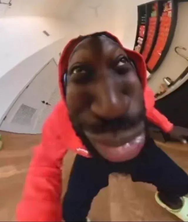 This image captures a person in a red hoodie and dark pants, rendered with an extreme fisheye lens effect that comically distorts their features. Their face, particularly the nose and mouth, appears exaggeratedly large and round, while their eyes are rolled upwards, conveying a humorous, almost bewildered or playful demeanor. The wide-angle perspective makes the subject seem to lean into the camera, creating an immersive, meme-like quality. The background, also warped by the lens, reveals an indoor setting with light-colored walls, a wooden floor, and what appears to be a display of red items on the right, possibly a collection or game setup. The entire scene exudes a lighthearted, internet-culture vibe, characteristic of viral comedic content rather than a specific show or event.