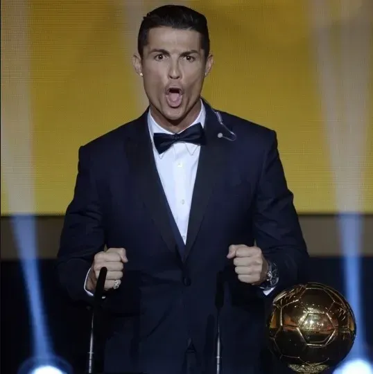 Cristiano Ronaldo is captured mid-roar, delivering his iconic "Siu" celebration with intense fervor on what appears to be an awards stage. Dressed impeccably in a black tuxedo, white shirt, and bow tie, his mouth is wide open, eyes wide, and fists clenched, embodying pure triumph. The golden Ballon d'Or trophy sits prominently beside him, confirming the prestigious event. The background features a warm, golden-yellow hue, illuminated by stage lights, perfectly complementing the golden trophy and the celebratory atmosphere of the Ballon d'Or ceremony. His dramatic roar, usually reserved for the pitch, is humorously amplified in this formal setting, making it seem like he's trying to summon the golden football itself to life.