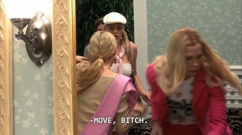 This chaotic image captures a humorous and confrontational moment from the movie 'White Chicks.' A blonde woman in a vibrant pink jacket, with her hair dramatically flying, appears to be forcefully pushing past another woman with a blonde ponytail and a pink shawl. The woman in the ponytail is reflected in an ornate gold mirror, looking back with a surprised or annoyed expression, while another woman in a white cap peers over her shoulder with wide eyes. The text overlay, '-MOVE, BITCH,' perfectly encapsulates the urgent and unrefined interaction. The background features elegant light blue floral wallpaper and a classic wall sconce, creating a comedic juxtaposition with the boisterous scene unfolding.