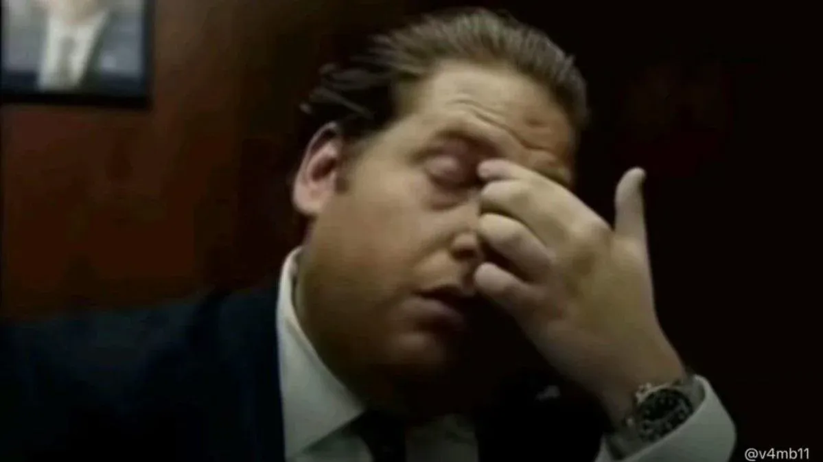 The image captures actor Jonah Hill, portraying his character Donnie Azoff from the film 'The Wolf of Wall Street.' He is seen in a close-up, wearing a suit and tie, with a look of profound exasperation or distress. His right hand is pressed against his closed right eye, as if rubbing away a headache or trying to process an overwhelming situation. The dark, indistinct background, possibly an office, adds to the somber mood, emphasizing his internal struggle. This particular still is a widely recognized meme, humorously depicting moments of extreme frustration, regret, or disbelief, perfectly capturing the chaotic and often absurd world of the film.