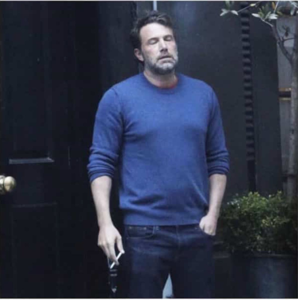 The image captures actor Ben Affleck in what has become the iconic 'Sad Affleck' meme. He stands outdoors, eyes closed and head tilted back slightly, sporting a weary and dejected expression, complete with a scruffy beard. He's dressed casually in a blue long-sleeved sweater and dark jeans, holding a cigarette in his left hand, with his right hand in his pocket. The background features a dark, possibly black, door with a brass doorknob on the left, and some sparse greenery, including a small tree and a potted plant, on the right, suggesting an urban or residential setting. The humor stems from his visibly exhausted demeanor, which has been widely adopted as a meme to convey feelings of disappointment, existential dread, or general weariness, often in response to life's mundane or frustrating moments.
