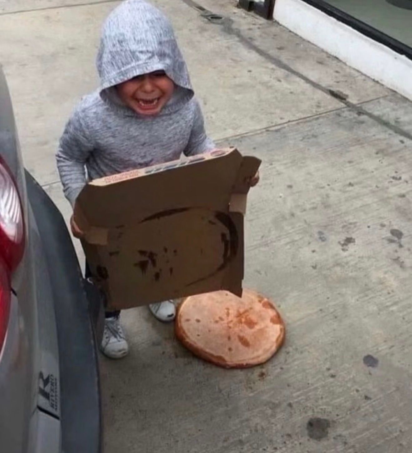 The image perfectly encapsulates the iconic "Crying Kid with Dropped Pizza" meme, showcasing a young child in a grey hoodie consumed by profound despair. Their face is contorted in a loud, tearful cry, eyes squeezed shut, as they clutch an empty pizza box. The source of their heartbreak, a freshly dropped pizza, lies tragically face-down on the grimy concrete ground next to a silver car. The mundane outdoor setting, with a hint of a building in the background, underscores the sudden and public nature of this culinary catastrophe. The humor lies in the universally relatable, yet comically exaggerated, devastation over a lost meal, a moment of pure, unadulterated childhood sorrow that resonates deeply.
