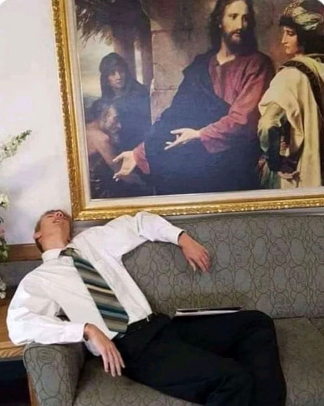 A young man, formally dressed in a white shirt and striped tie, is completely sprawled and seemingly asleep on a patterned grey sofa. His head is tilted back, mouth slightly open, conveying utter exhaustion or perhaps boredom. Directly above him hangs a large, ornate gold-framed painting depicting a religious scene, likely Jesus Christ, with an outstretched hand. The humorous juxtaposition is striking: the serene, spiritual figure in the painting appears to be gesturing directly towards the deeply slumbering man below, almost as if offering a blessing for his nap or questioning his attentiveness. The formal setting, suggested by the attire and artwork, makes the man's relaxed demeanor even funnier. I did not find any famous or recognizable content in this image.