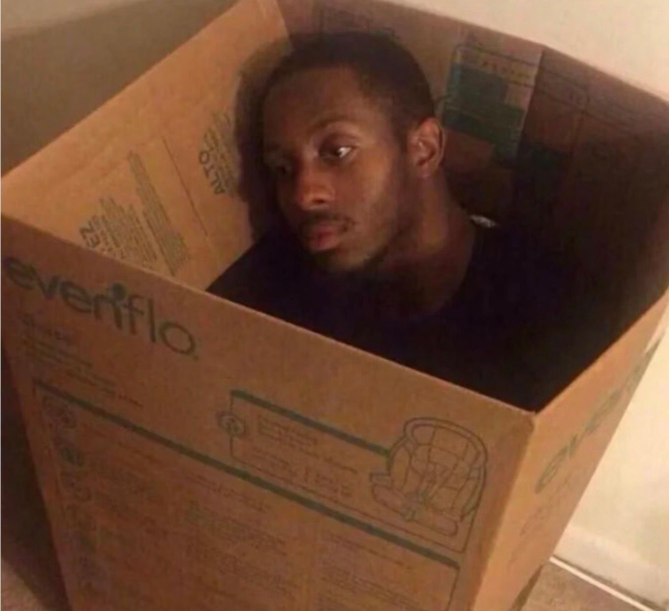 This image captures a man, known from the popular 'man in a box' meme, peering out from inside a large cardboard box. His expression is a mix of bewilderment and mild resignation, with his eyes looking slightly upwards and to the side, conveying a sense of being trapped or perhaps just observing his surroundings from an unusual vantage point. The box itself is prominently branded with 'Evenflo', a company known for baby products like car seats, which adds a layer of humorous irony to an adult occupying it. The background is simply the interior of the box, emphasizing his confinement. The humor stems from the absurdity of an adult in a child's product box, perfectly encapsulating the meme's essence of feeling stuck or out of place.