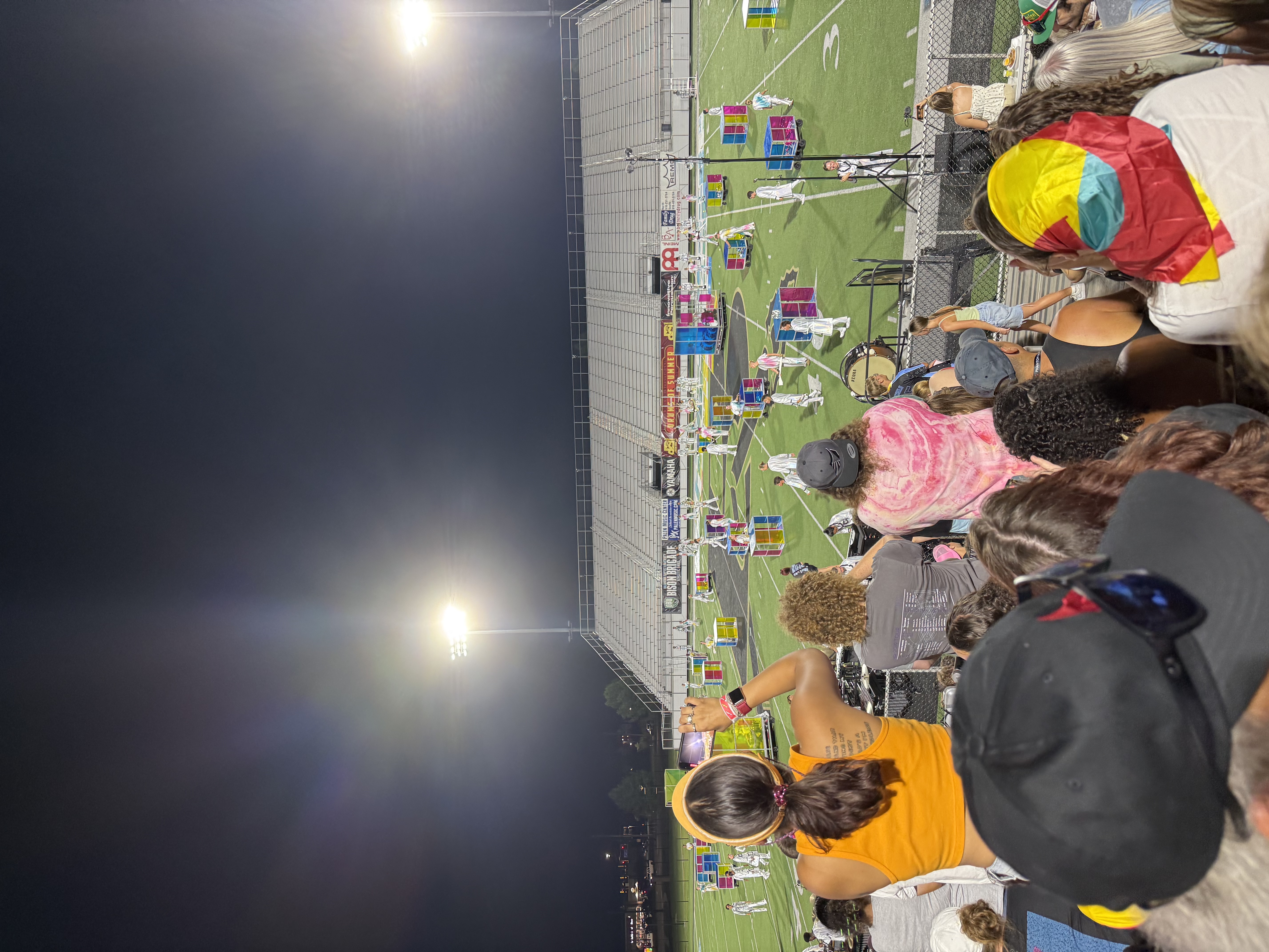 The image captures a lively scene at a nighttime event, likely a performance or competition on a sports field, illuminated by bright stadium lights. In the foreground, a diverse crowd watches intently, some spectators with colorful attire, indicating a festive atmosphere. The central focus appears to be on performers in white outfits engaging in a structured activity, surrounded by vibrant, multicolored props scattered across the field.
The background features the stadium bleachers, filled with spectators, which adds to the sense of community and excitement. The performers seem to be part of a choreographed routine, possibly a marching band or color guard, enhancing the event's dynamic energy.
Humorously, one might imagine the performers discussing their strategies, like “Should we go for a synchronized pirouette or a dramatic pose?” while the audience enthusiastically debates who has the best dance moves! The overall scene radiates fun and celebration, typical of a lively community event.