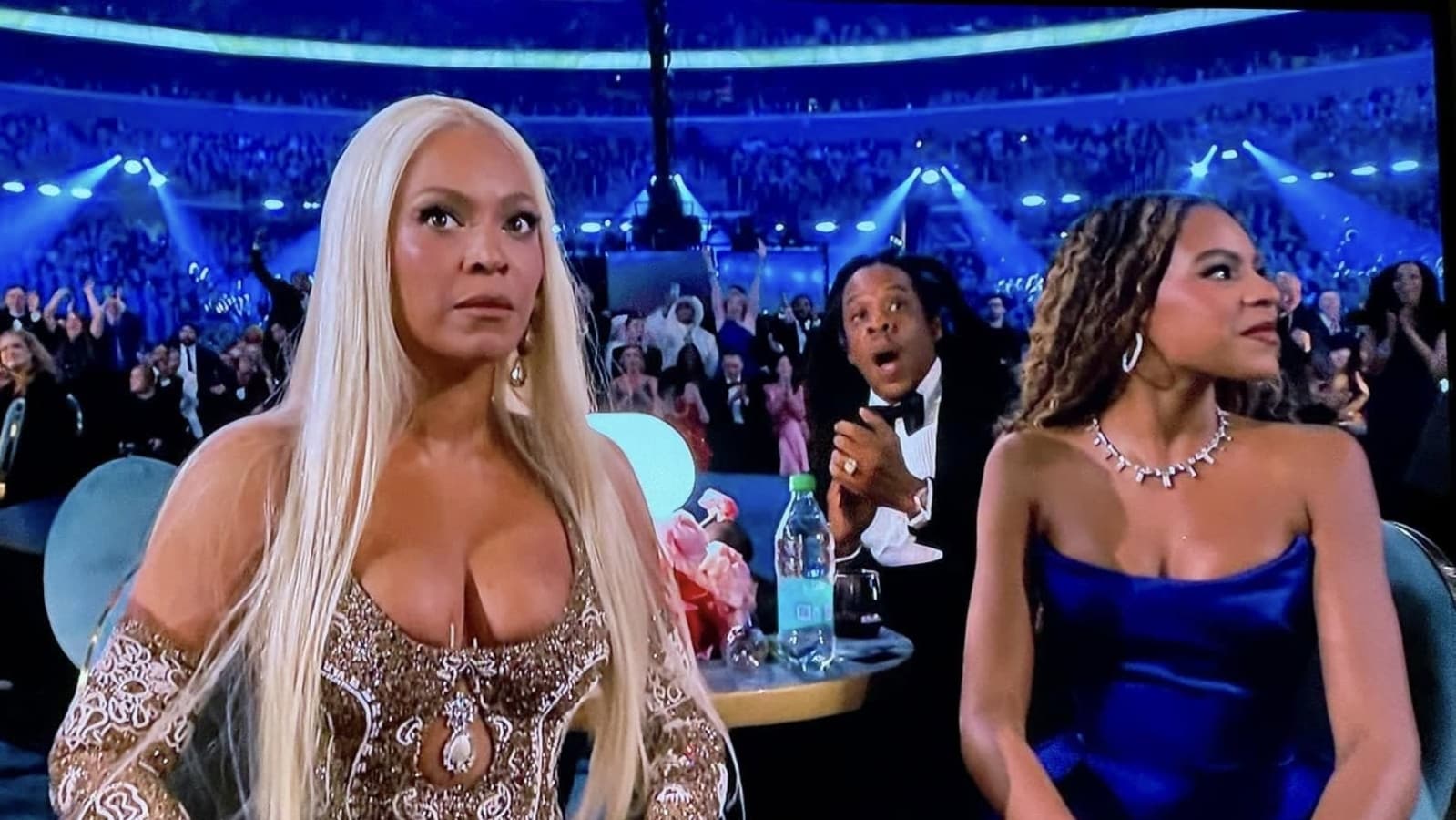 In this image from the Oscars, Beyoncé, dressed in a stunning embellished gown, displays a serious, almost perplexed demeanor, while seated next to Jay-Z, who appears animated and surprised. Their expressions suggest they are reacting to a significant moment on stage, possibly a shocking win or performance. 

To their right, a woman in a blue dress looks on with a subtle smile, indicating a mix of emotions in the atmosphere. The background is filled with a cheering crowd, bathed in blue lights, emphasizing the high-energy environment of the awards ceremony. 

The contrast between Beyoncé's composed demeanor and Jay-Z's expressive surprise adds a layer of humor, as if he's witnessing something unbelievable, while she remains the epitome of poise. This snapshot encapsulates the blend of glamour and genuine reactions that makes such events memorable.
