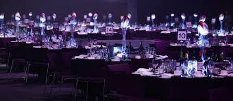 The image depicts a lavish banquet setup with numerous elegantly dressed tables, each adorned with stylish centerpieces. The mood is both anticipatory and festive, suggesting a significant event, perhaps a gala or awards ceremony. Each table is meticulously arranged with glimmering glassware and neatly folded napkins, indicating a high level of detail and sophistication.
The background is dimly lit, enhancing the focus on the tables while casting a soft glow on the decor. The numbers on the tables imply an organized seating arrangement, possibly for an event where guests will be honored or celebrated.
The atmosphere is likely filled with excitement, as guests await the festivities. Humorously, one might imagine the tables whispering secrets about the guests they’re about to host, each hoping to steal the spotlight with their dazzling decor!