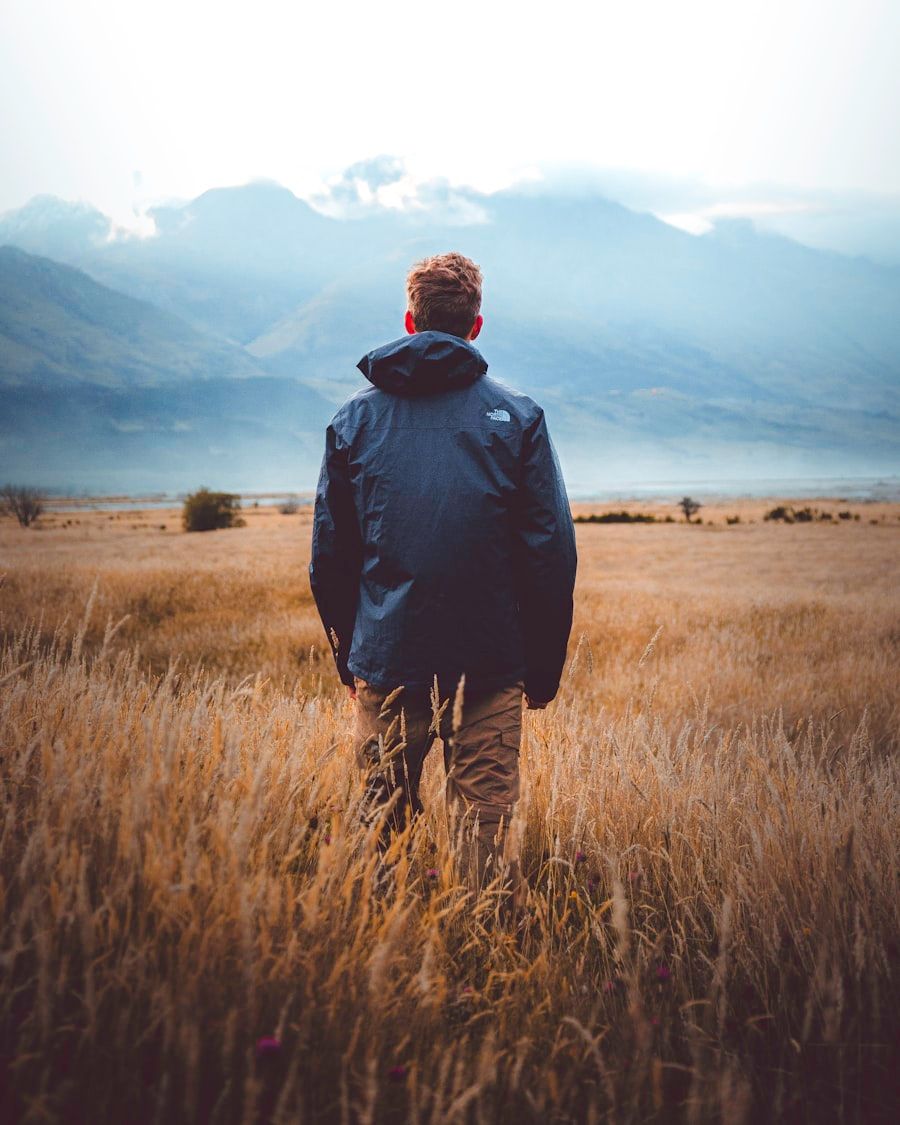 The subject stands with their back to the viewer, gazing towards distant, cloud-shrouded mountains across a vast field of golden grass. Their posture is relaxed, shoulders slightly dropped, head subtly tilted upwards, indicating a contemplative focus on the expansive landscape. Dressed in a practical dark blue North Face jacket and khaki cargo pants, their attire signals readiness for outdoor elements and an affinity for nature. This clothing choice projects an image of an adventurous, self-reliant individual. The solitary stance in this grand, open environment suggests a desire for introspection, peace, or perhaps a quiet sense of awe. There's no apparent discrepancy between what is projecteda connection with nature and a spirit of explorationand what is communicated through their absorbed, unposed presence. The scene evokes a feeling of quiet solitude and immersion in the natural world.