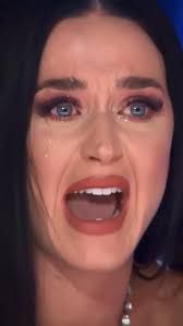 The image captures a close-up of Katy Perry, her face contorted in an intense expression of distress and sadness. Her mouth is wide open as if mid-wail, revealing her teeth, and her blue eyes are wide and tearful, with visible tears streaming down her cheeks. Her dark hair frames her face, and she wears dramatic eye makeup, which appears slightly smudged from her tears. The background is dark and out of focus, emphasizing her raw emotion. This iconic image is widely recognized as a popular meme, often used humorously to exaggerate feelings of disappointment or despair, particularly stemming from her time as a judge on 'American Idol' during an emotional moment. The humor lies in the sheer intensity of her expression, making it relatable yet comically over-the-top.