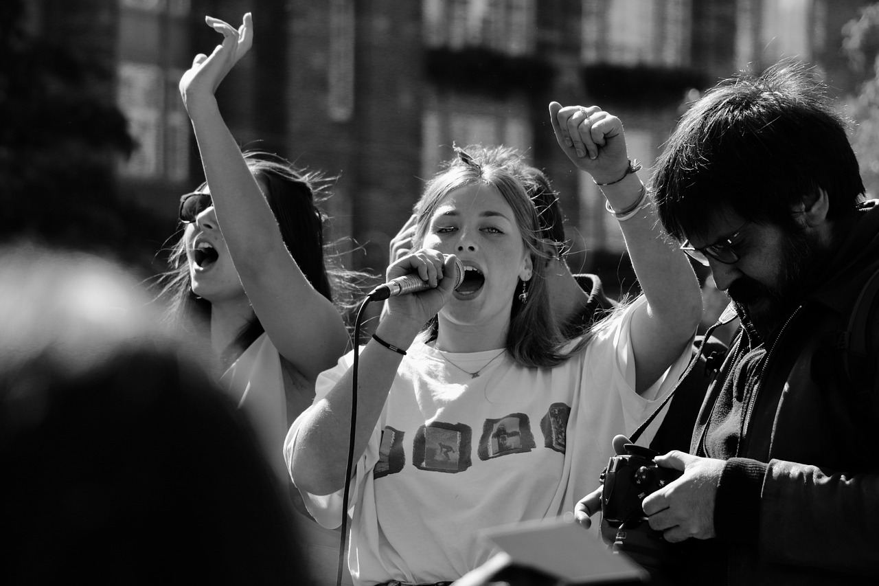 In this striking black-and-white image, a passionate young woman stands at the forefront, microphone in hand, her mouth wide open as she delivers an emphatic message. Her raised arm and clenched fist suggest empowerment and urgency, indicative of a protest or rally. She wears a casual T-shirt adorned with graphic prints, which adds a youthful vibe to her assertive stance. 

Behind her, another figure appears to support the rally, while a man with a camera captures the moment, highlighting the event's significance. The blurred background hints at a crowd, suggesting a collective effort and shared purpose. The atmosphere is charged with energy, embodying the spirit of activism. 

The image humorously contrasts the intensity of the moment with the relaxed attire of the participants, like they’re ready to both rally and chill at a café afterward!