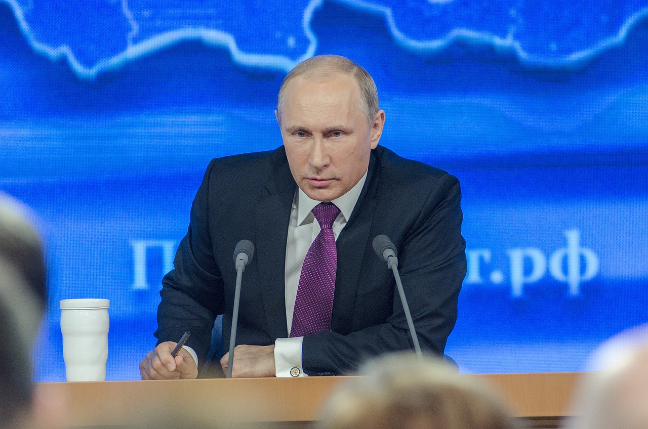 Vladimir Putin is captured in a formal setting, likely a press conference or official address, seated at a wooden table with two microphones in front of him. He wears a dark suit, a crisp white shirt, and a distinctive purple patterned tie. His demeanor is serious and focused, with a direct gaze that conveys intensity. To his left, a plain white travel mug adds a touch of everyday reality to the otherwise formal scene. The background is a large, vibrant blue screen displaying a subtle map-like graphic and partial Cyrillic text, possibly a Russian domain like ".рф", reinforcing the official Russian context of the event. The blue backdrop provides a professional yet somewhat stark environment, highlighting Putin as the central figure. One might humorously imagine him pondering the profound implications of his coffee choice amidst global affairs.