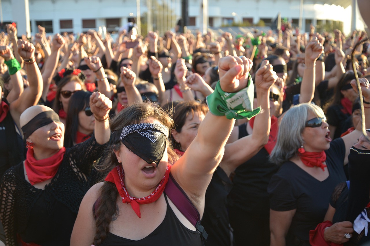 In this vibrant image, a large group of women is engaged in a passionate protest, their fists raised high in solidarity. Many participants are wearing blindfolds and bandanas, suggesting a unified movement advocating for social or political change. Their expressions convey determination and fervor, with some shouting passionately, while others chant in unison.
The background features a crowd that stretches far, indicating a significant turnout for this demonstration. The sunlight casts a warm glow, emphasizing the energy in the scene. The coordinated use of black clothing, red bandanas, and green accessories hints at a specific cause, though the exact context isn't clear.
Humorously, one could imagine the women synchronizing not just their attire but their battle cries, as if rehearsing for an epic protest performance. Each fist raised could be seen as a dramatic punctuation mark in their rallying cry for justice!