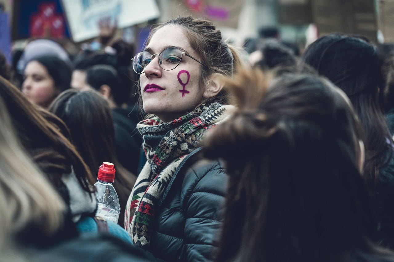A young woman with a resolute expression and round glasses is captured in a close-up, her face painted with a bold purple female symbol, indicating her participation in a protest. Her dark pink lipstick and patterned scarf add a touch of personal flair to her serious demeanor. She wears a practical puffer jacket, suggesting a chilly outdoor event. Her gaze is directed upwards, conveying a focused and determined spirit amidst the bustling crowd. The blurry background of other attendees and faint signs confirms a large public gathering. A water bottle peeking into the frame humorously reminds us that even the most passionate activists need to stay hydrated, proving that revolution, much like life, requires proper fluid intake.